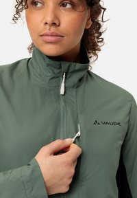 Giacca leggera verde con colletto alto, chiusura frontale con zip e tasche laterali. Presenta il logo nero "VAUDE" sul petto. Texture morbida.