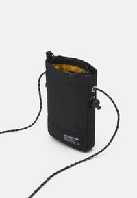 Sac en tissu noir avec fermeture à cordon, doté d'un intérieur jaune. Comprend une étiquette de marque et une sangle réglable à motifs.