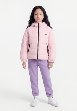 Kind mit langen dunklen Haaren, das eine hellrosa Steppjacke, lila Jogginghose und weiße High-Top-Sneaker trägt, steht vor weißem Hintergrund.