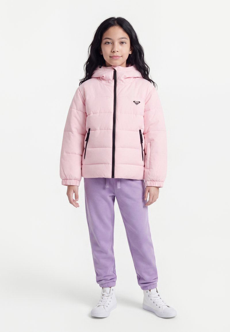 Enfant aux longs cheveux foncés portant une doudoune rose clair, un pantalon de jogging violet et des baskets montantes blanches, debout sur fond blanc.