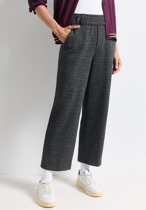 Personne portant un pantalon large gris foncé, des chaussettes blanches, des baskets blanches et une veste bordeaux, les mains dans les poches.