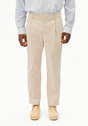 Homme debout, portant une chemise à carreaux bleu clair, un pantalon plissé beige et des mocassins beiges, sur un fond uni.