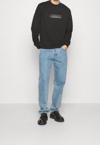 Felpa nera con logo stampato, jeans azzurri chiari e scarpe nere con suola spessa. Il tessuto appare liscio; i jeans hanno una vestibilità classica.