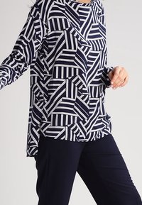 Marinblå långärmad tröja med ett geometriskt vitt randigt mönster. Löst sittande med rund halsringning och en asymmetrisk nederkant.