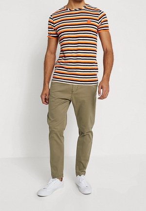 Stripet t-shirt med sorte, orange og hvide linjer; rund halsudskæring; parret med lyse khakibukser og hvide sneakers.