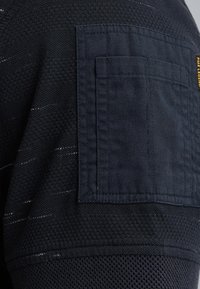 Tumma laivasilkkinen tekstiili, jossa on neulekuvio ja neliömäinen läpällinen tasku. Erityispiirteinä on kontrastivärinen ompelu ja sileä helma.