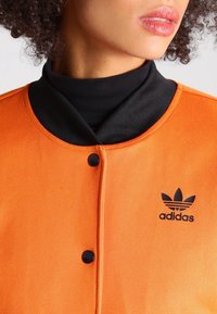 Orange jacka med svart krage, med knappstängning och en svart Adidas-logotyp. Texturerad tyg med en slät finish.