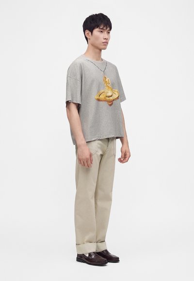 Νέος άνδρας στέκεται φορώντας γκρι oversized μπλουζάκι με κίτρινο γραφικό, μπεζ παντελόνια και σκουρόκαφέ loafers σε απλό λευκό φόντο.