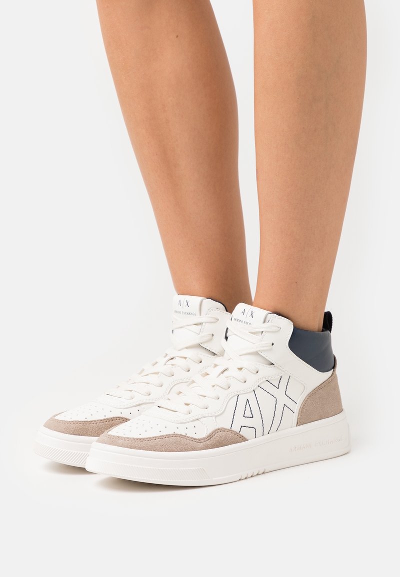Baskets montantes blanches avec des accents en daim beige et un détail de col noir portées aux pieds sur fond uni.