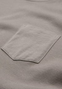 Tessuto grigio chiaro con una trama a costine e una piccola tasca sul petto. Il materiale è morbido con cuciture visibili lungo i bordi.