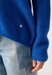 Blue knitted sweater featuring a ribbed texture, side slits, and a silver logo button. Paired with light blue jeans showcasing a faded pattern.