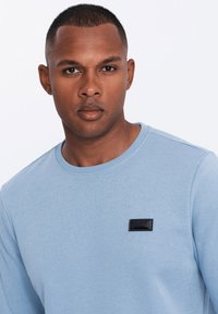 Sudadera azul claro con cuello redondo y puños acanalados, que presenta un detalle de etiqueta rectangular negra en la parte delantera.