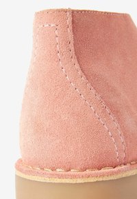 Botte de cheville en suède rose avec des coutures beiges, forme arrondie, texture douce et semelle extérieure de couleur claire.