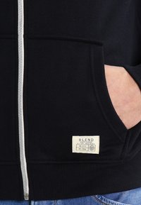 Hauts à zip noir avec une poche kangourou, des poignets côtelés et une étiquette tissée à l'ourlet, fabriqué à partir d'un tissu doux pour le confort.