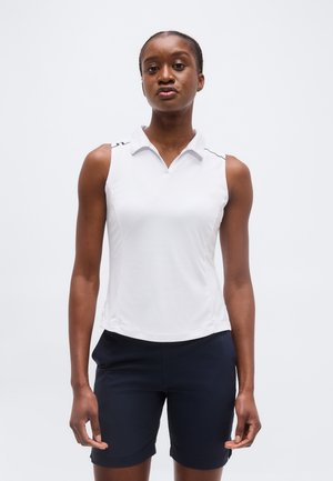 Γυναίκα που φοράει λευκό πόλο χωρίς μανίκια και μαύρο σορτς, στέκεται μπροστά σε απλό λευκό φόντο.