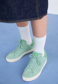 Mint green suede sneakers with matching accents, white textured socks, and a white rubber sole, positioned on a blue surface.