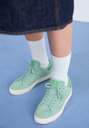 Baskets en daim vert menthe avec accents assortis, chaussettes blanches texturées et semelle en caoutchouc blanche, posées sur une surface bleue.