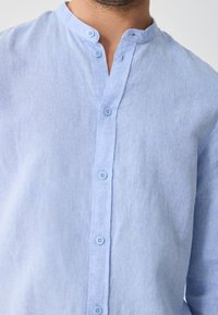 Light blue linen shirt featuring a mandarin collar, front button closure, and subtle texture. Light buttons complement the fabric's hue.
