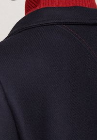 Marineblauer Mantelkragen mit diagonalem Muster, akzentuiert durch rote Nähte. Darunter ist ein mehrlagiger roter Rippenpullover sichtbar.