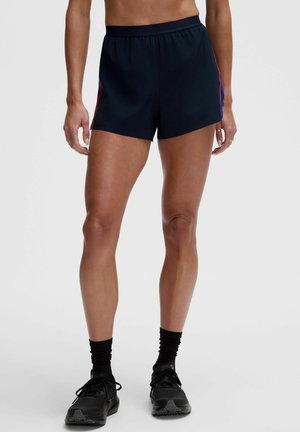 Frau trägt schwarze Sportshorts mit roten Seitenstreifen, schwarze Socken und schwarze Laufschuhe, steht vor einem einfarbigen Hintergrund.