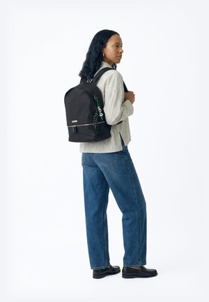 Femme portant une chemise rayée, un jean bleu et des mocassins noirs, avec un sac à dos noir orné d'un porte-clés décoratif sur une épaule.