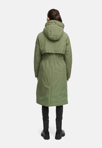 Olijfgroen lange parka met een trekkoord in de taille, een capuchon en geborduurde merknaam op de achterkant. Draagt over zwarte broeken en laarzen.