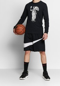 Person wearing black long-sleeve shirt with classical figure graphic, black and white shorts, black Nike socks, and black sneakers holding a basketball.