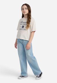 T-shirt grafico beige chiaro con maniche corte e stampa blu navy, abbinato a jeans azzurri a gamba larga e sneaker nere basse.