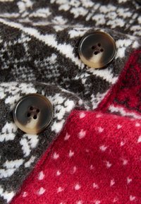 Neulottu villakangas, jossa on tumma kuvio valkoisilla koristeilla, täydentävänä punaiset osiot ja kaksi suurta ruskeaa nappia.