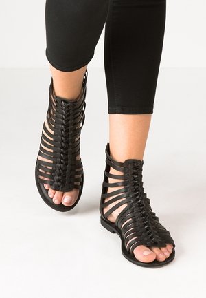 Czarne sandały na paski z odkrytymi palcami, ozdobione plecionymi detalami i pionowymi paskami, wykonane z elastycznego materiału z płaską podeszwą.