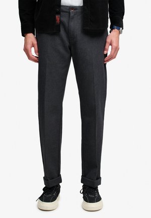 Hombre con pantalones gris oscuro de pierna recta remangados en los puños, zapatillas negras con suelas blancas y una chaqueta negra sobre una camisa blanca.