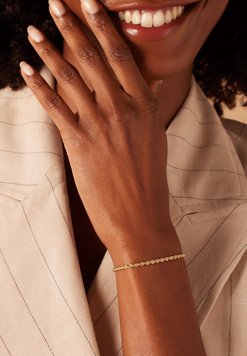 Gold braided bracelet worn on a bare wrist, displayed against a light-colored, pinstriped fabric background. Smooth texture and delicate design.