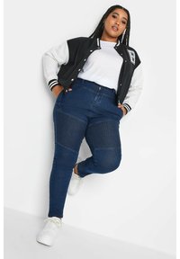 Jeans ajustados azul oscuro con paneles de rodilla texturizados, combinados con una chaqueta varsity negra y blanca sobre una camiseta blanca y zapatillas blancas.