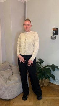 Bež pletena jopa s kablovskim vzorcem, zlati gumbi, nošena preko belega vrha. Črne široke hlače in črni čevlji. Notranja scena s rastlinami.