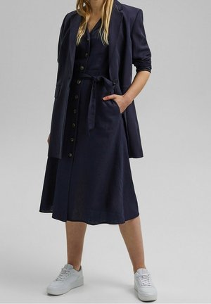 Marineblauwe linnen jurk met een nauwsluitend lijfje, knoopsluiting aan de voorkant en een gestrikte taille. Gecombineerd met een bijpassende blazer en witte sneakers.