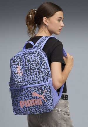 Jongere draagt een zwart shirt en grijze spijkerbroek, en heeft een paarse en zwarte rugzak met patroon van Puma met een roze logo.