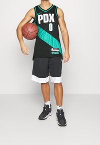 Maillot de basket-ball noir avec accents verts, logo "PDX" et numéro "0". Associé à un short noir avec finitions blanches et des baskets noires.