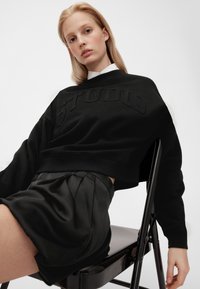 Sudadera negra recortada con texto en relieve "STUDIO", combinada con una falda plisada de satén negra. Modelo sentado en una silla negra, fondo neutro.