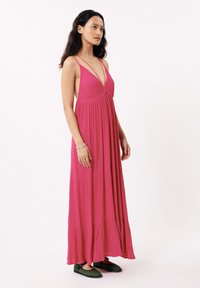 Roze maxi-jurk met spaghettibandjes, vloeiende stof en gelaagd ontwerp. Model draagt groene schoenen met een zwarte zool en een zilveren armband.