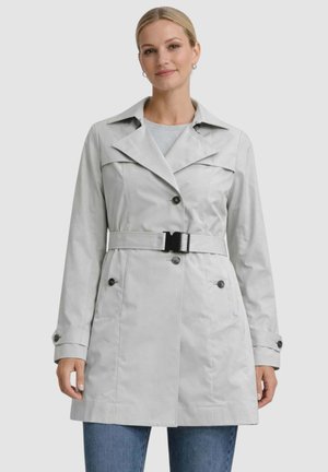 Donna che indossa un trench leggero grigio chiaro con cintura, collo ampio, polsini con bottoni e tasche frontali, abbinato a jeans blu.