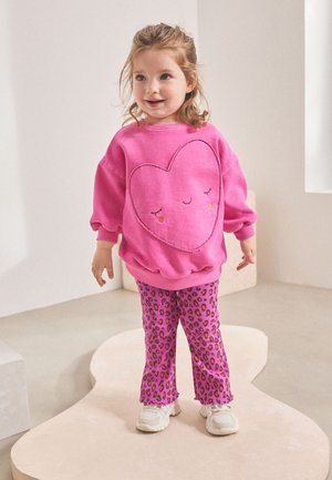 Junges Mädchen steht drinnen und trägt ein leuchtend pinkes Sweatshirt mit einem lächelnden Herz und rosa Leopardenhose.