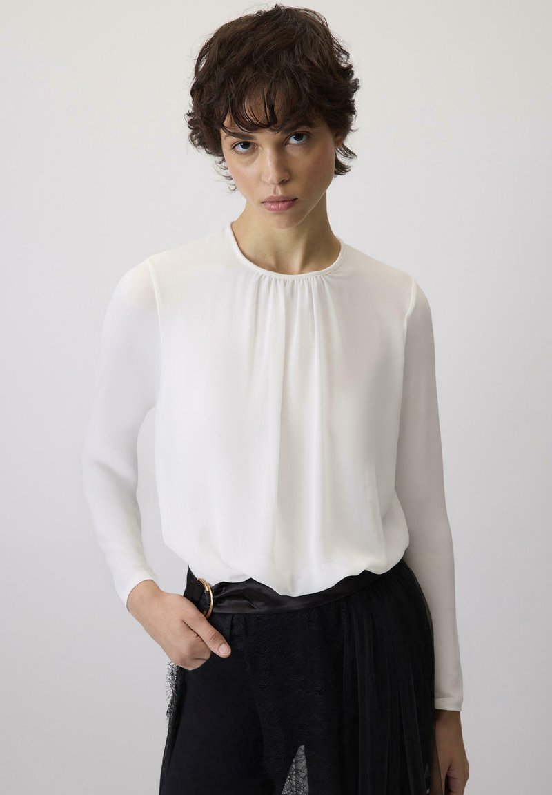 Witte blouse met lange mouwen en een geplooide halslijn, gemaakt van gladde, lichtgewicht stof. Gecombineerd met een zwarte riem en een geplooide zwarte rok.