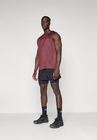 Weinrotes ärmelloses Sportoberteil mit strukturiertem Muster, kombiniert mit schwarzen Shorts und schwarzen Sportschuhen, steht vor einem neutralen Hintergrund.