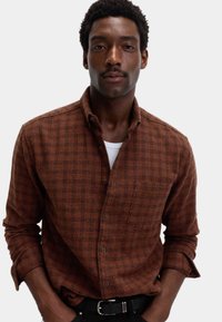 Camisa de cuadros marrón con botones, cuello estructurado, bolsillo en el pecho y mangas enrolladas, hecha de una tela tejida texturizada.