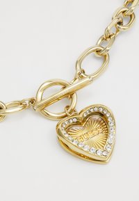 Bracciale color oro con grandi anelli intrecciati, caratterizzato da un ciondolo a forma di cuore decorato con cristalli trasparenti e la scritta "GUESS".