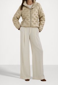 Beżowa pikowana kurtka z wysokim kołnierzem, dwiema przednimi kieszeniami i zamkiem błyskawicznym. W połączeniu z kremowymi spodniami o szerokich nogawkach i brązowymi butami.