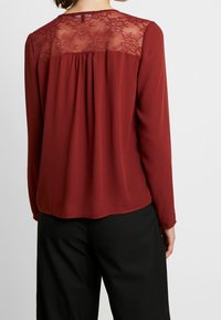 Blouse bordeaux à manches longues, panneau arrière en dentelle transparente, détail froncé à la taille, texture lisse et ourlet arrondi.