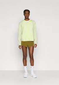 Ελαφρύ, ημιδιαφανές κίτρινο μπουφάν με ρυθμιζόμενο καπό, φερμουάρ και ελαστικά μανίκια. Συνδυάζεται με ελιά σορτς και λευκά αθλητικά παπούτσια.