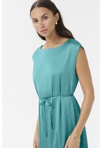 Une robe sans manches de couleur bleu-vert présente un tissu lisse et texturé ainsi qu'un détail de taille à nouer. L'encolure a une forme douce bateau.