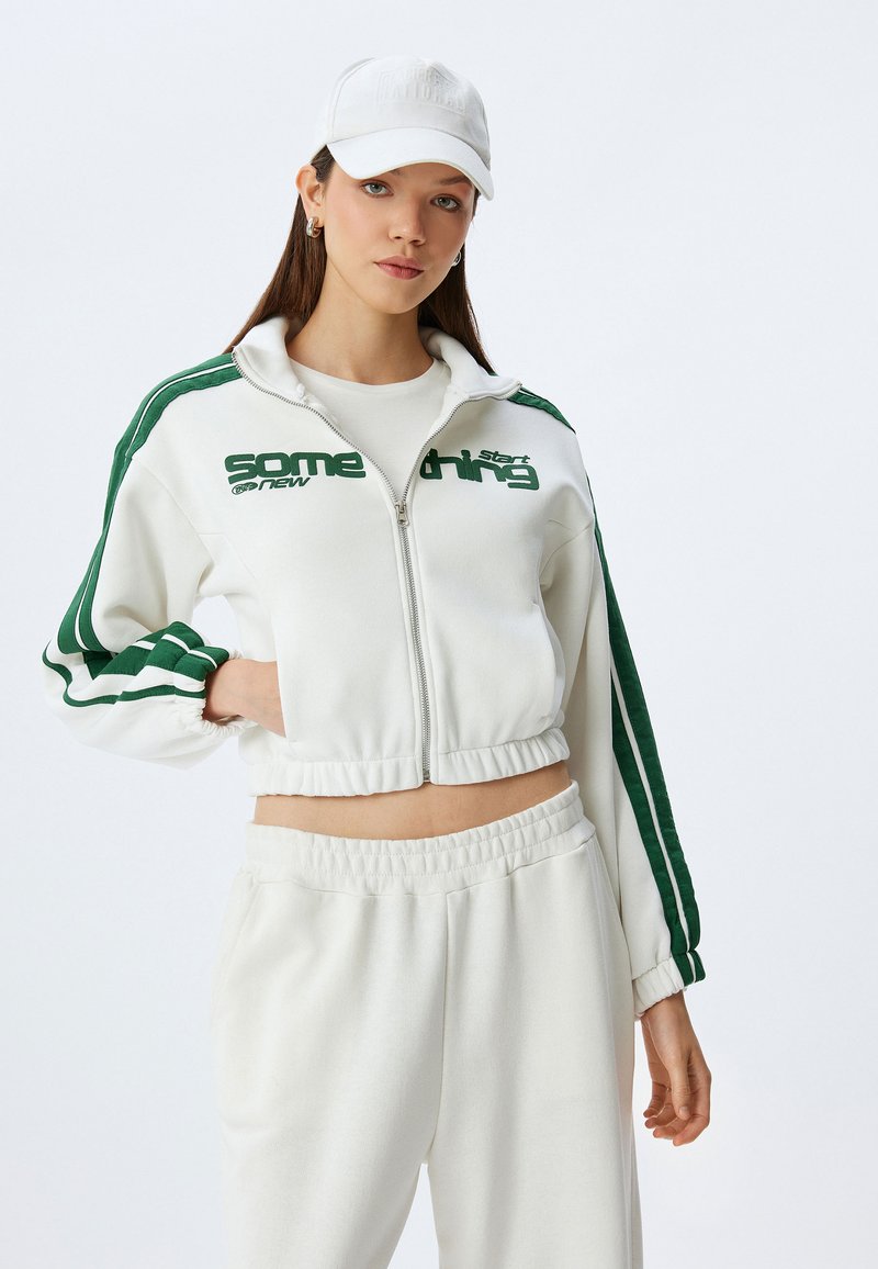 Witte cropped zip-up jas met groene strepen, voorzien van tekstgraphics. Gecombineerd met bijpassende witte sweatpants. Een witte pet maakt de outfit compleet.